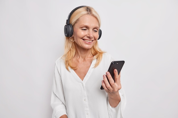 Felice positiva bionda di mezza età bella donna ha videoconferenza tiene il telefono cellulare focalizzato sullo schermo dello smartphone utilizza cuffie wireless indossa camicetta bianca pone al coperto