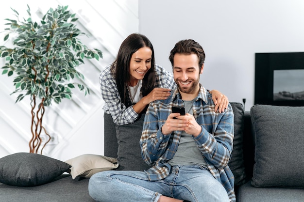 Happy positive caucasian spouses in a trendy casual clothes\
spend leisure time at home together in living room use the phone\
they watch the news browse social networks choose home goods\
smile