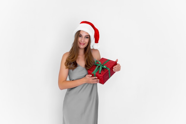 Happy positive girl in santas hat holds christmas gift while stands on white background celebration