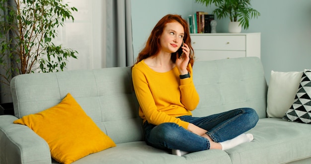 Счастливая и позитивная девушка сидит на диване у себя дома и говорить на своем смартфоне