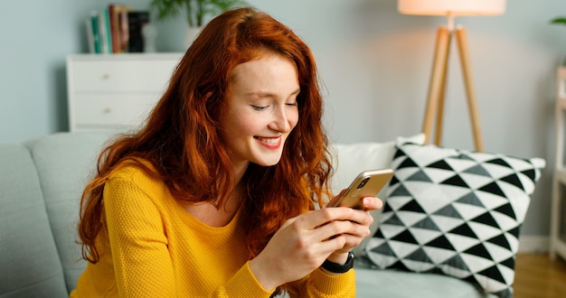 Happy and positive girl sitting on sofa at home and using smartphone