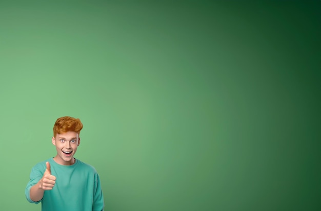 Happy positive smiling attractive redheaded teenage guy with curly hair looking at camera wearing casual trendy tshirt standing isolated on green background over studio copy space background