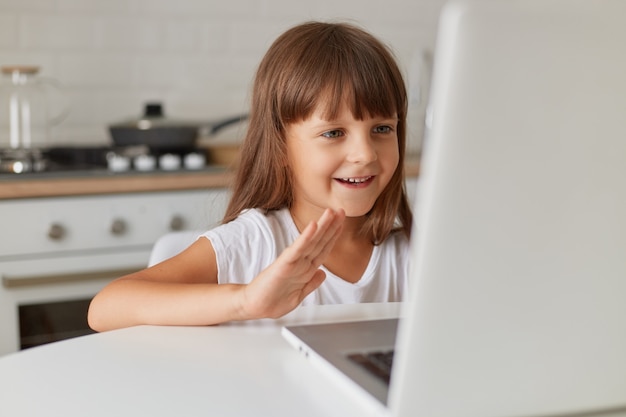 Happy positive smiling female kid with dark hair sitting in kitchen in front of laptop computer, having video call or broadcasting livestream in her child blog.