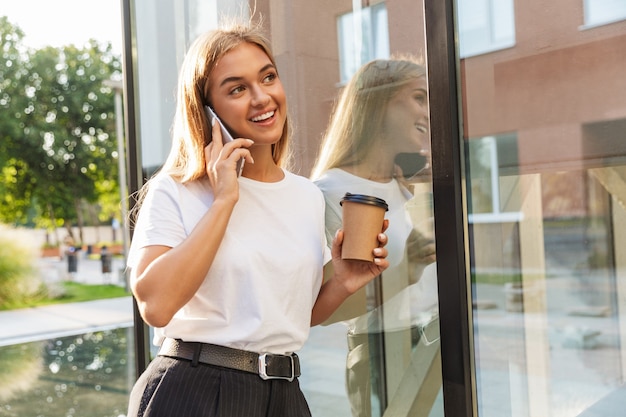 Giovane donna sorridente positiva felice di affari con la tazza di caffè che posa all'aperto vicino al centro di affari che parla dal telefono cellulare.