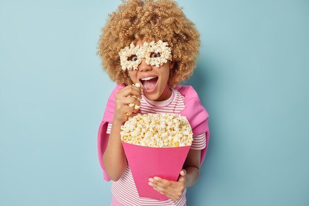 Happy positive woman with curly hair eats tasty popcorn watches\
movie film in cinema makes eyemask of corn dressed in casual\
clothes poses over blue background enjoys leisure time home\
entertainment