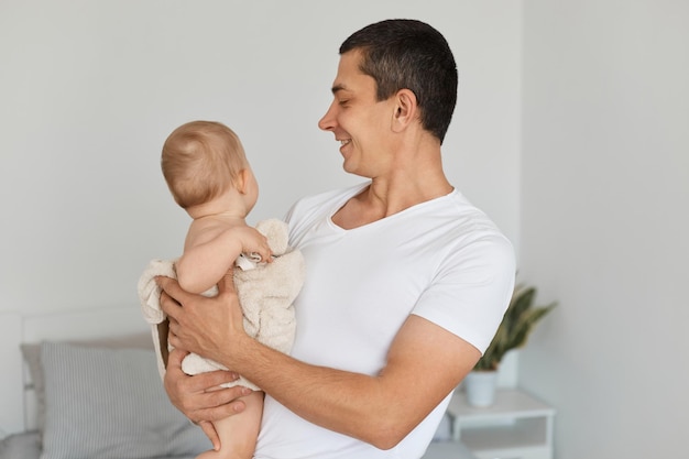 幸せなポジティブな若い大人の父親と彼の幼児の女の赤ちゃんが家で一緒に遊んで、笑顔で彼の娘を見て、愛を表現し、子供の世話を楽しんでいます。