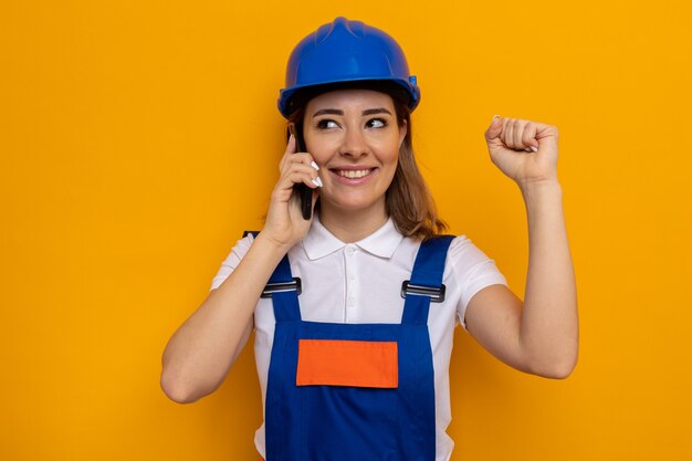 Happy and positive young builder woman in construction uniform and safety helmet clenching fist while talking on mobile phone