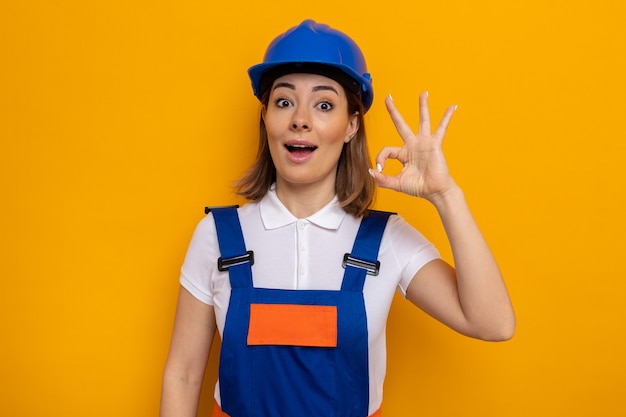 Happy and positive young builder woman in construction uniform and safety helmet looking smiling doing ok sign