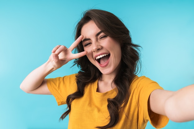 happy positive young pretty woman posing isolated over blue wall take a selfie by camera showing peace gesture.