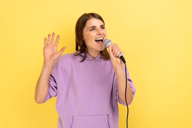 Happy positive young woman standing holding microphone singing songs in karaoke with raised arm, having fun alone, wearing purple hoodie. Indoor studio shot isolated on yellow background.