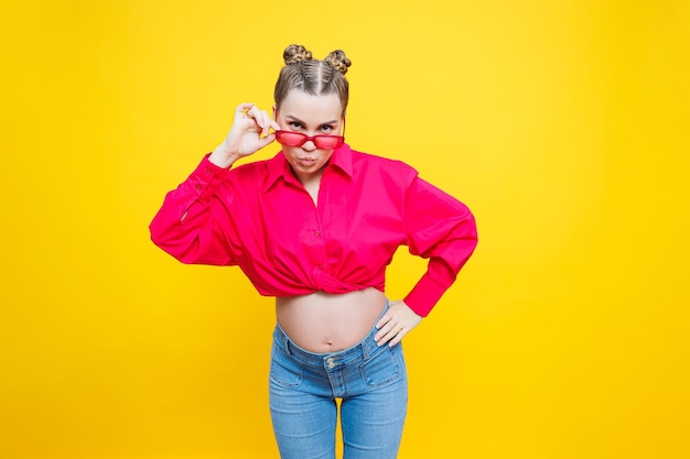 Happy pregnancy Cheerful pretty pregnant woman in a pink shirt and pink glasses on a yellow background Young bright pregnant woman