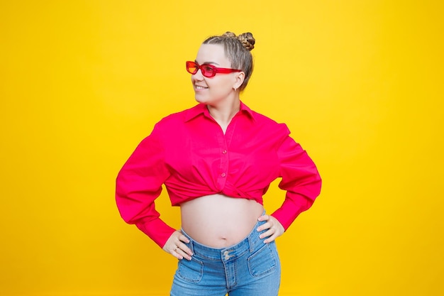 Happy pregnancy Cheerful pretty pregnant woman in a pink shirt and pink glasses on a yellow background Young bright pregnant woman