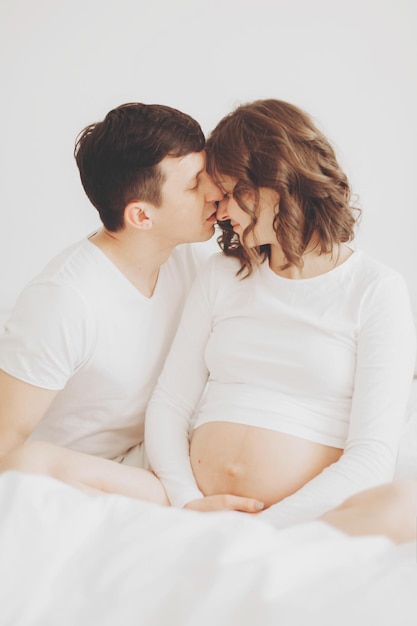 Happy pregnant couple relaxing on white bed and holding belly\
bump happy young husband kissing his smiling wife and hugging baby\
belly stylish pregnant family in white at home true happiness