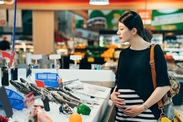 Happy pregnant female customer choosing and buying fish in shop. young future parenthood lady holding big belly with baby inside doing grocery shopping in supermarket looking at fresh seafood.