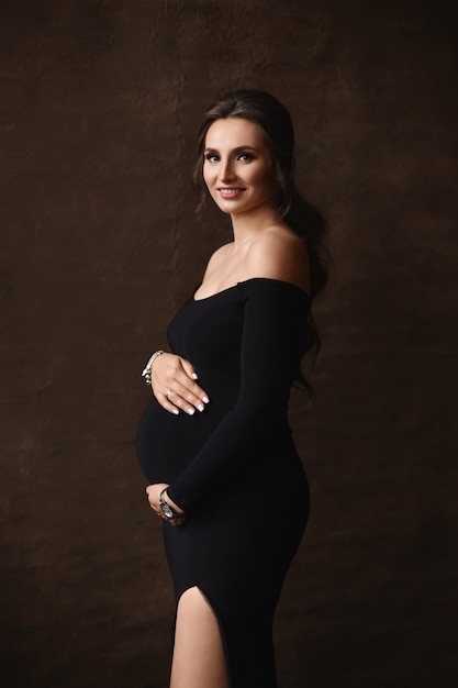 A happy pregnant girl wearing a trendy evening dress holds her belly and poses on a brown background. Young pregnant woman in long black dress posing over brown background.