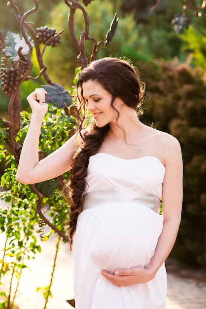 A happy pregnant girl in a white dress is walking in the park.