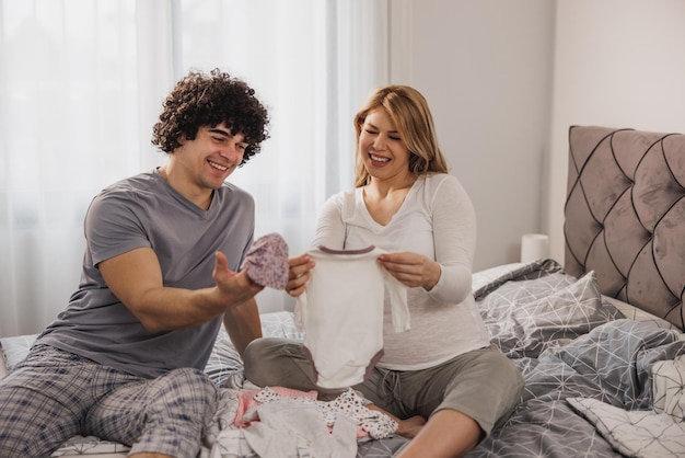 Happy pregnant mother showing baby bodysuit to husband at home.