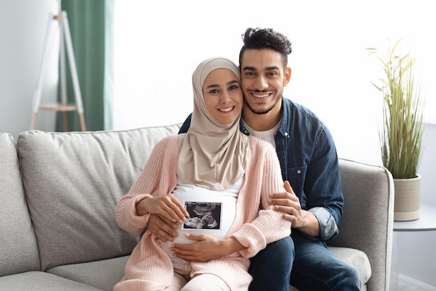 Happy Pregnant Muslim Family Sitting On Couch And Holding Baby Sonogram Picture