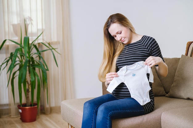 Happy pregnant woman in casual clothes sitting on sofa and holding little dress