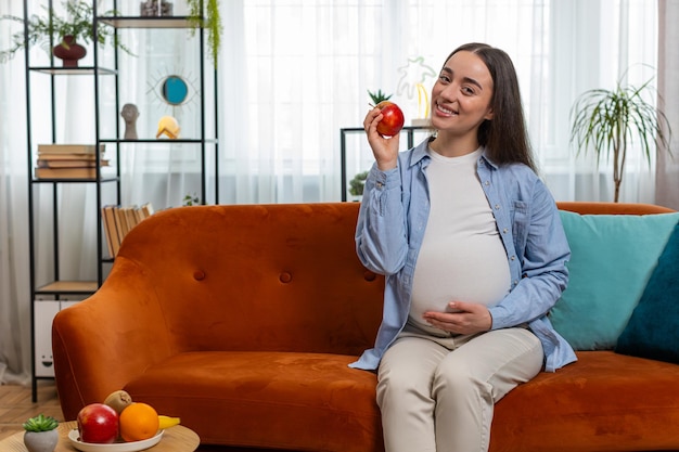 happy pregnant woman eating fresh red apple fruit sitting on sofa healthy vitamin food motherhood
