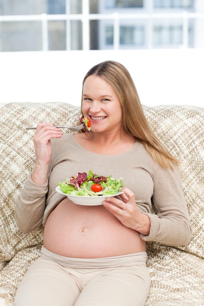 Happy pregnant woman enjoying a salad sitting on the sofa