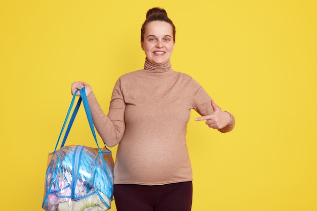 Happy pregnant woman pointing at her stomach, smiling expectant mother being ready going to maternity hospital, looks happy, dressed casually, posing isolated over yellow wall.