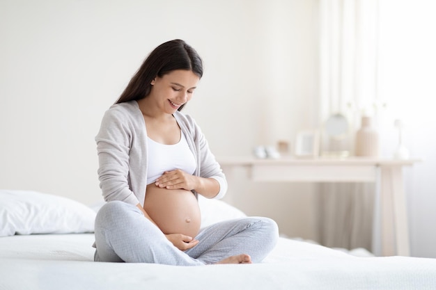 彼女の寝室のベッドの上に座って幸せな妊娠中の女性