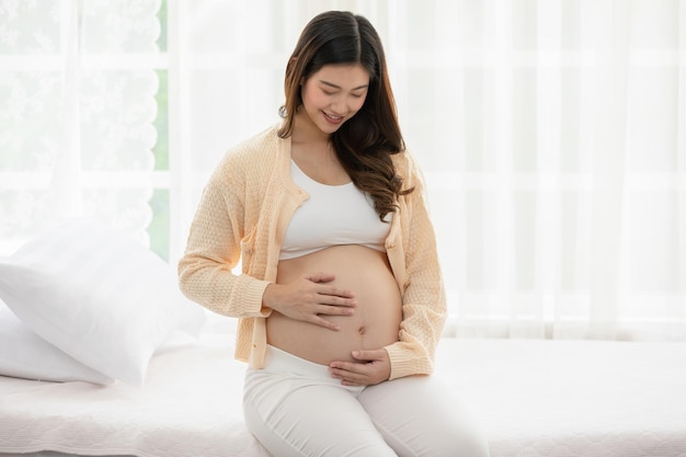 행복한 임신한 여성은 침대에 앉아 집에서 큰 배를 들고 만지고 젊은 여성의 임신은 미래의 삶을 즐기고 집에서 편안하게 휴식을 취합니다 모성 및 임신 개념 부드러운 초점