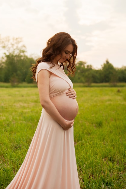 Happy pregnant woman walking in the green field in park at sunrise 