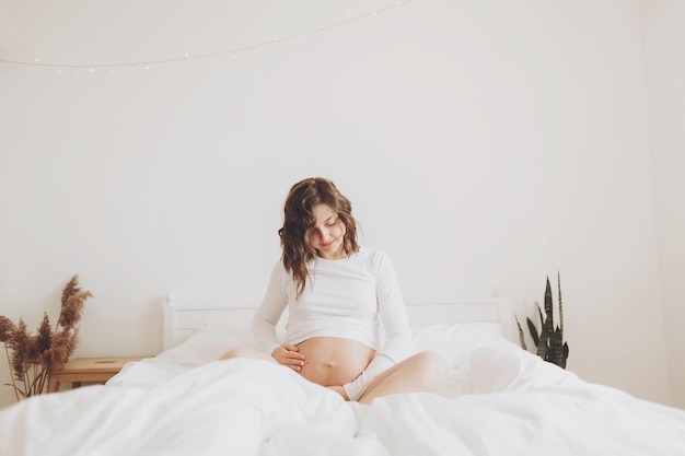 Счастливая беременная женщина в белом держит живот и расслабляется на белой кровати дома Стильная беременная мама ждет ребенка Концепция материнства и фертильности Время материнства