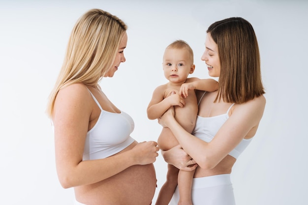 Happy pregnant woman with big belly and beautiful mother with baby on colored background -  Young women wearing underwear expecting a baby - Pregnancy, motherhood, people and expectation concept