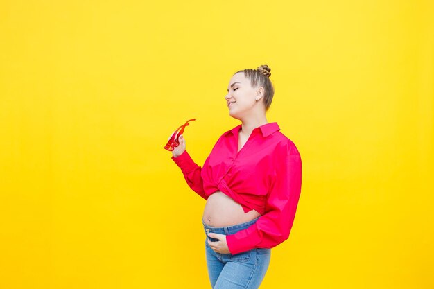 Happy pregnant woman with a smile on her face Cheerful pretty pregnant woman in a pink shirt and pink glasses on a yellow background Young bright pregnant woman