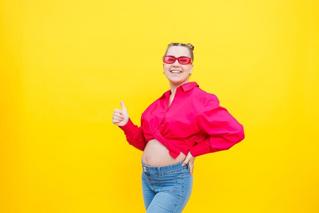 Happy pregnant woman with a smile on her face Cheerful pretty pregnant woman in a pink shirt and pink glasses on a yellow background Young bright pregnant woman