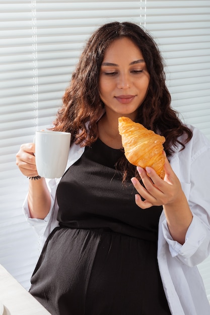 幸せな妊娠中の若い美しい女性は、朝の朝食時にクロワッサンを食べてブラインドを探しています。妊娠中の心地よい朝と前向きな姿勢