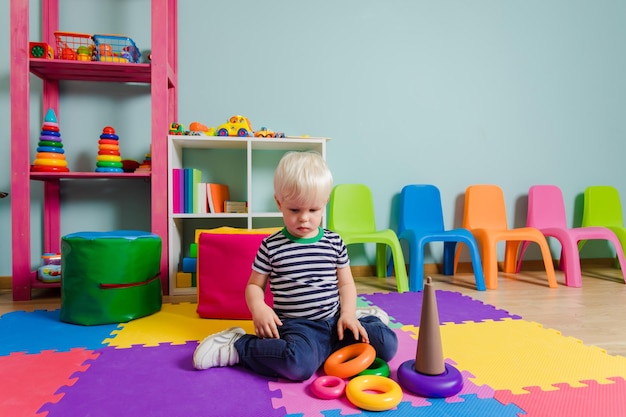 幸せな就学前の男の子がプレイルームで遊んでいます彼は床に座っており、彼の隣にはピラミッドの色付きのリングが散らばっています