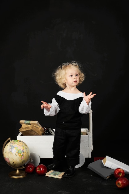 Happy preschool girl near the cart with a books, globe and clock on a black background 