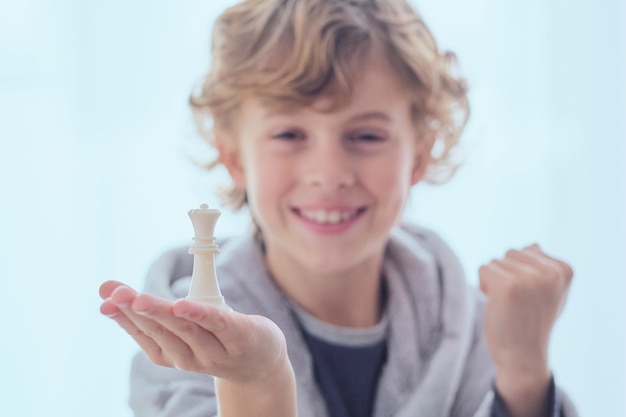 Счастливый мальчик-подросток демонстрирует шахматную фигуру ферзя и поднимает кулак, радуясь победе