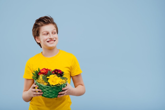 彼の手に花のバスケット、青い壁に分離された黄色のTシャツ、コピースペース、目をそらして幸せなプレティーンの少年