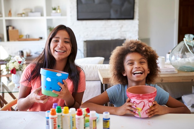 Happy preteen girl and her older girlfriend holding plant pots that theyave decorated with paints at home smiling to camera close up