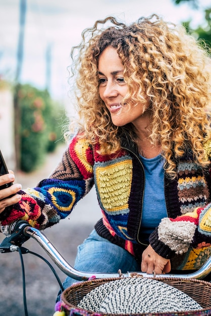 Happy pretty adult young woman enjoy outdoor leisure activity on a bike and read cell phone smiling woman people use modern cellular to send message blonde long curly hair cheerful middle age lady