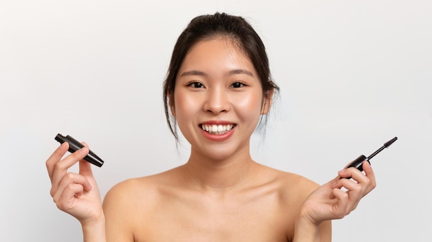 마스카라를 들고 흰색 스튜디오 배경에서 포즈를 취하는 카메라를 보며 웃고 있는 행복한 예쁜 아시아 여성