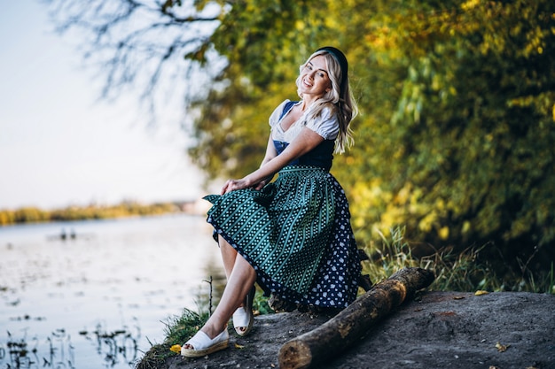 Happy pretty blond girl in dirndl, traditional beer festival dress, sitting outdoors with blured colorful trees behind