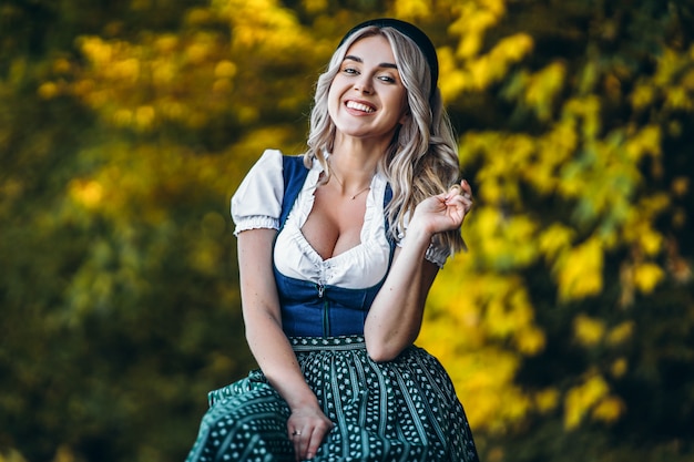 Ragazza bionda graziosa felice in dirndl, vestito tradizionale da festival della birra, sedentesi all'aperto con gli alberi colourful blured dietro