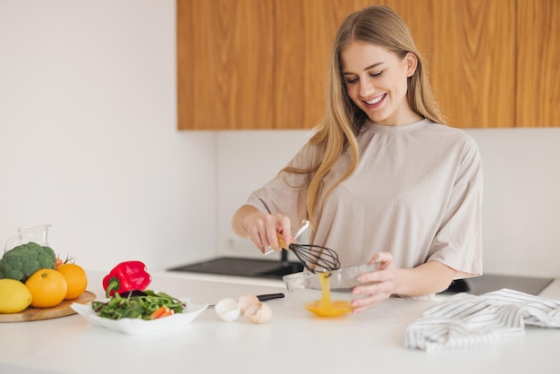 パジャマを着た幸せなかなり金髪の女性が、自宅のキッチンで卵と新鮮な野菜の朝食を準備しています