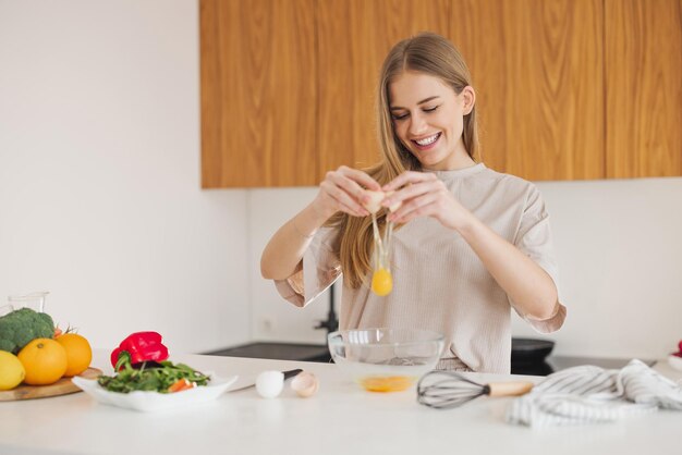 Happy pretty blonde woman in pajamas is preparing breakfast of eggs and fresh vegetables in kitchen