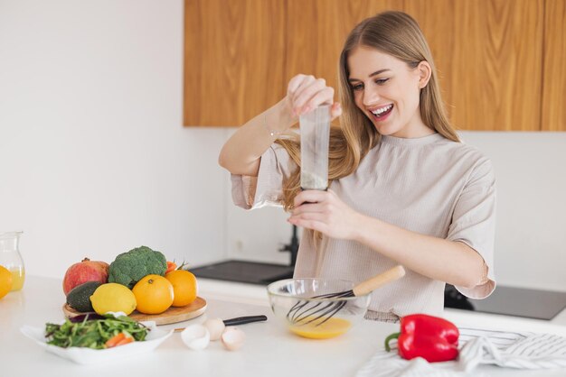 パジャマを着た幸せな金髪の女性が、自宅のキッチンで新鮮な野菜の中で卵と塩の朝食を準備している