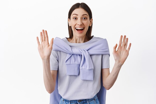 Happy pretty female model raising hands up, showing empty palms and looking excited at front, waving to say hello, smiling friendly, standing over white wall surprised