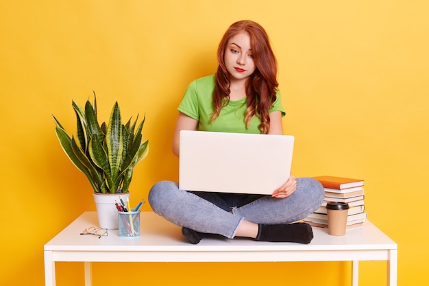 Happy pretty ginger woman sitting on table and working with laptop