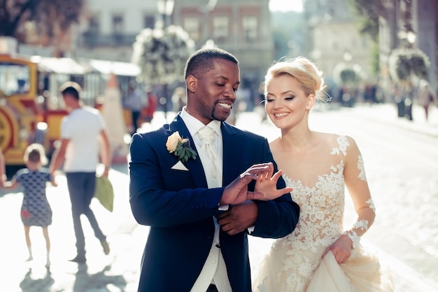 Happy pretty girl or cute bride with beautiful blond hair in white sexy wedding dress and handsome man or african American groom walking outdoors on sunny day on blurred city street background