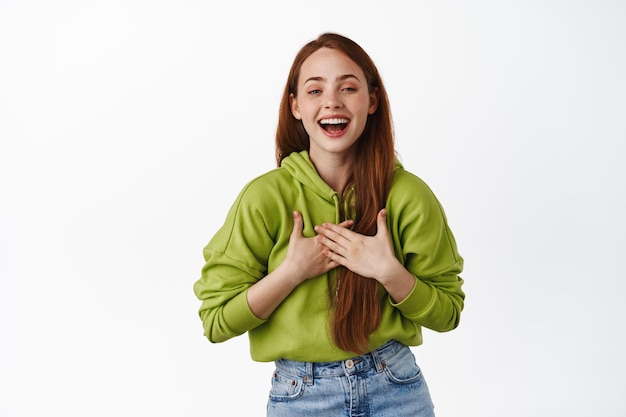 Happy pretty girl laughing, holds hands on heart and smiling pleased, say thank you, looking joyful and delighted, standing over white background.